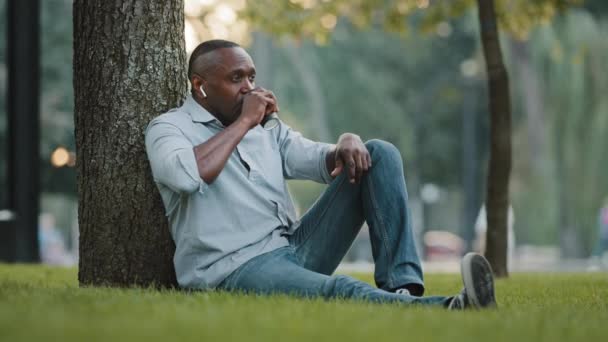 Пожилой африканский бизнесмен-предприниматель, сидящий на траве, отдыхающий рядом с деревом в городском парке во время перерыва. Взрослый человек наслаждается ароматным горячим кофе или чаем в одноразовой чашке на открытом воздухе - Кадры, видео