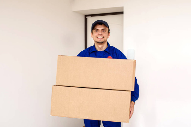 Jonge vrolijke glimlachende mannenkoerier in blauwe overall bezorgt een pakket in twee kartonnen dozen. Witte achtergrond met deur - Foto, afbeelding