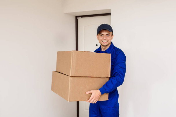 Junger glücklich lächelnder Mann in blauem Overall bringt ein Paket in zwei Kartons. Weißer Hintergrund mit Tür - Foto, Bild