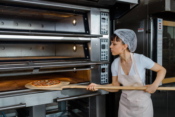 Młoda biała kobieta piekarz trzyma skórkę drewna ze świeżą pizzą i wkłada ją do piekarnika w piekarni. Smaczna koncepcja fabryki pizzy. - Zdjęcie, obraz