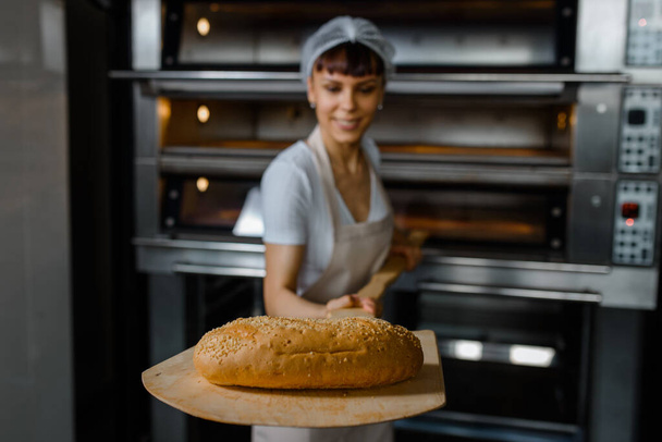 Young caucasian woman baker is holding a wood peel with fresh bread near an oven at baking manufacture factory. Tasty bread bakery factory concept. Selective focus on a bread - Photo, Image