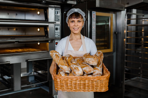 Молодая кавказская пекарь держит в руках большую корзину свежего хлеба на хлебопекарной фабрике. Фон с электрическими печами. Концепция хлебной пекарни. Счастливая улыбающаяся женщина смотрит в камеру - Фото, изображение