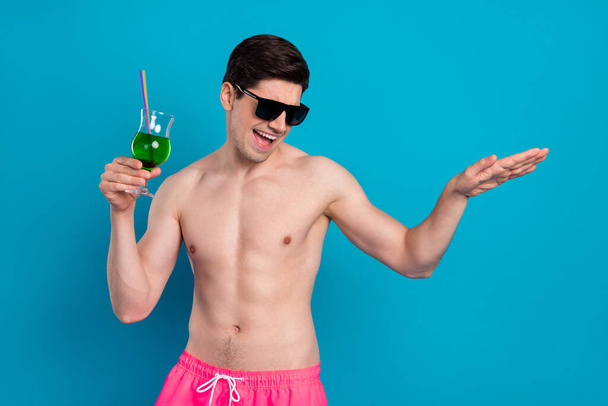 Foto von lustigen charmanten jungen Mann nackten Oberkörper dunkle Brille hält Cocktail tanzen lächelnd isoliert blaue Farbe Hintergrund - Foto, Bild
