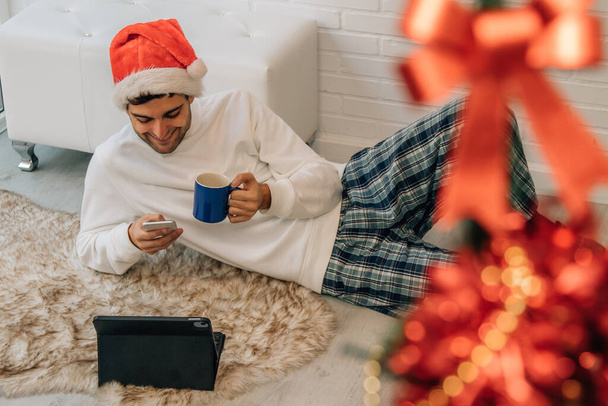 мужчина в шляпе Санта Клауса дома с мобильным телефоном и компьютером - Фото, изображение