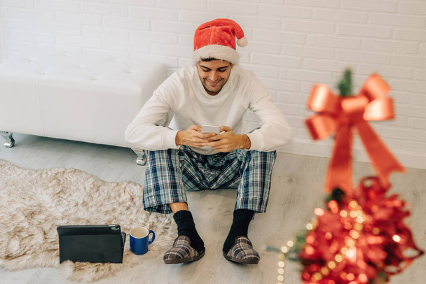 Mann mit Weihnachtsmannmütze zu Hause mit Handy und Computer - Foto, Bild