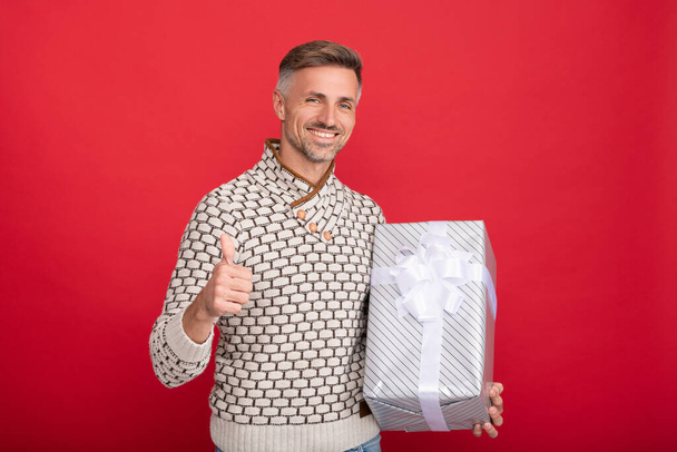 счастливый мужчина в свитере держит подарочную коробку на красном фоне, показывая большой палец вверх, день рождения - Фото, изображение