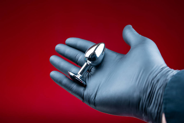Erotik bdsm oyunları için gümüş anal fiş tutan siyah eldivenli eller. - Fotoğraf, Görsel