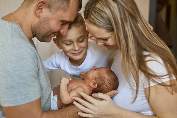 Father and mother holding newborn daughter in arms, older brother stands next to and looks with love. Family values - Zdjęcie, obraz