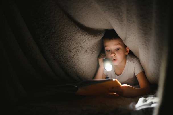 Reading book and using flashlight. Young boy in casual clothes lying down near tent at evening time. - Zdjęcie, obraz