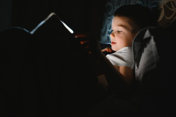 Portrait of cute little boy reading in bed with flashlight in dark room, enjoying fairytales - Foto, imagen