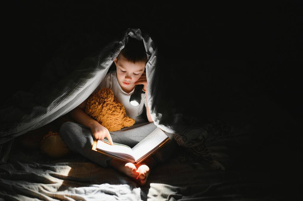 Boy with flashlight reading book under blanket at home - Foto, imagen