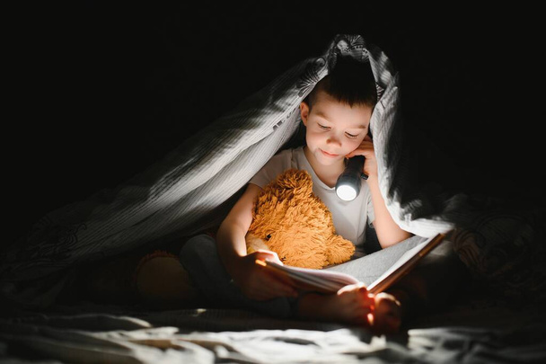 Child reading book in bed. Kids read at night. Little boy with fairy tale books in bedroom . Education for young children. Bedtime story in the evening. Cute kid under blanket in dark room with lamp. - Zdjęcie, obraz