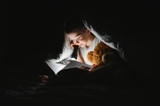 Boy with flashlight reading book under blanket at home - Foto, afbeelding