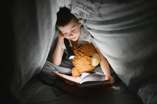 Reading book and using flashlight. Young boy in casual clothes lying down near tent at evening time. - Foto, immagini