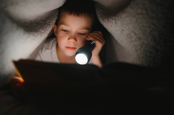 Boy with flashlight reading book under blanket at home - Foto, imagen