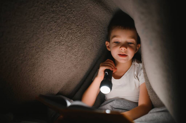 Boy with flashlight reading book under blanket at home - Photo, image