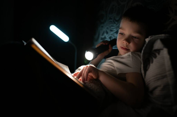 Lecture du livre et utilisation de la lampe de poche. Jeune garçon en vêtements décontractés couché près de la tente au moment du soir. - Photo, image