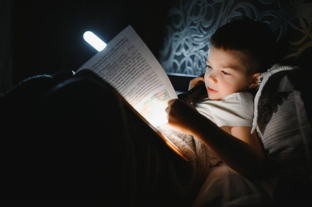 Child reading book in bed. Kids read at night. Little boy with fairy tale books in bedroom . Education for young children. Bedtime story in the evening. Cute kid under blanket in dark room with lamp. - Фото, изображение