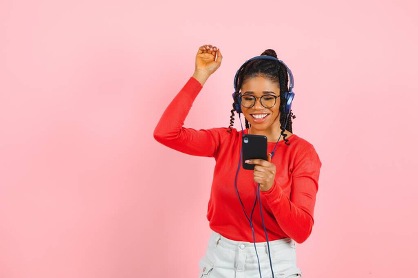 Pretty african american young woman with bright smile dressed in casual clothes, glasses and headphones dance over pink background. - Foto, afbeelding