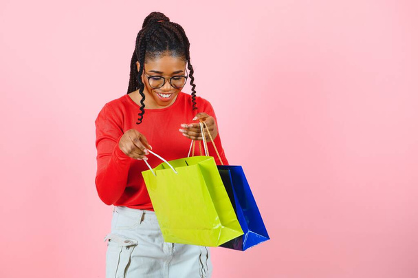 Velký prodej. Nadšený afro dívka s nákupními taškami dotýkající se slunečních brýlí, při pohledu na kameru přes růžové studio zeď - Fotografie, Obrázek