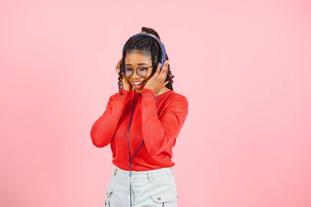 Mujer joven afroamericana bonita con sonrisa brillante vestida con ropa casual, gafas y auriculares bailan sobre fondo rosa
. - Foto, imagen