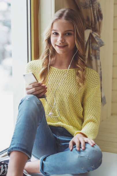 Прекрасна дівчина-підліток в навушниках за допомогою мобільного телефону, дивлячись на камеру і посміхаючись, сидячи на підвіконні
 - Фото, зображення