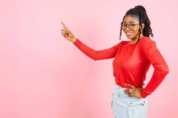 Young black african american smiling happy curly woman promoter 20s in blue shirt point index finger aside on copy space area mock up workspace isolated on pastel pink color background studio portrait - Foto, imagen