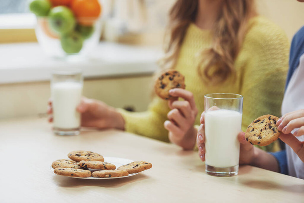 Lovely teenage girl and her beautiful mother eating cookies and drinking milk while sitting in the kitchen, close-up - Foto, afbeelding