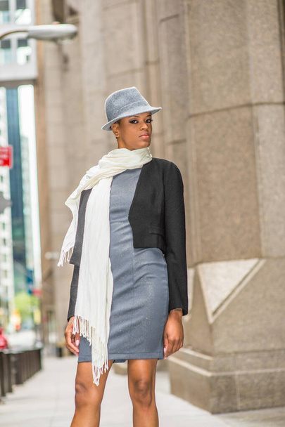Vestida com uma jaqueta preta de lã cortada, um vestido cinza, um longo lenço branco ao redor do pescoço, usando um chapéu Gangster Fedora de lã, uma mulher negra está de pé na rua - Foto, Imagem
