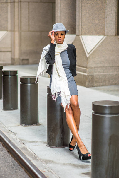 Dressing in a cropped jacket,  a fitted dress, open toes shoes and a long scarf, wearing a woolen Fedora hat, a young black woman is sitting on a metal column outside a building, relaxing and thinking - Fotografie, Obrázek