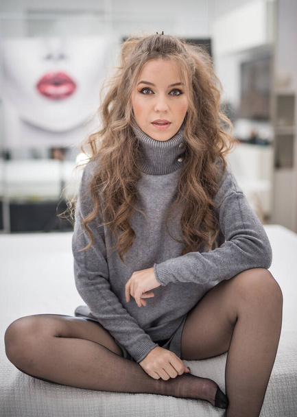 Attractive woman in black pantyhose and long curly hair posing challenging in  bed. Classic boudoir scene. Sensual young  woman with long curly hair and grey sweater looking at camera - 写真・画像