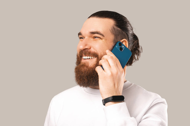 Photo of smiling young bearded man talking on smartphone over grey background - Valokuva, kuva