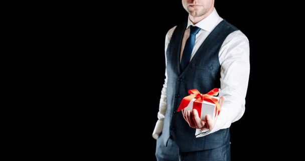 Caja de regalo de mano. Feliz joven hombre de negocios sosteniendo regalo sorpresa regalo con cinta roja aislada sobre fondo negro. San Valentín, Navidad, Año Nuevo banner con espacio para copiar - Foto, imagen