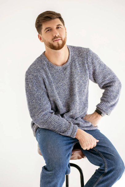 Handsome man with a beard on a white background in a photo studio. A confident man in denim trousers and a gray sweater sits on a chair. Vertical portrait of a 30 year old man - Foto, imagen