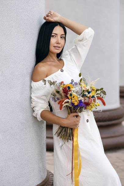 A beautiful young brunette girl in a white dress with a bouquet of wildflowers leaned against a column and poses. Vertical portrait of a cute girl with long black hair in a vintage or rustic style - Fotografie, Obrázek