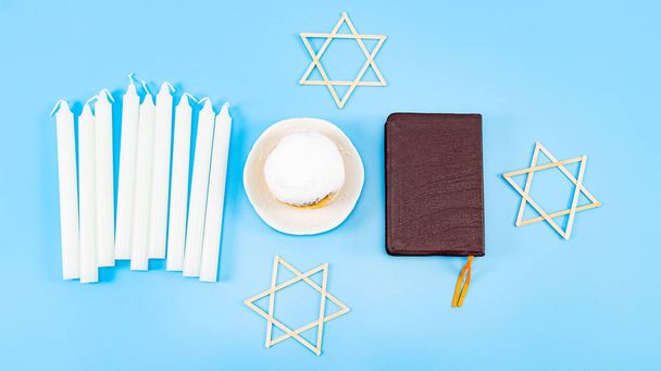 Ένα μεγάλο ντόνατς με σκόνη και κρέμα σε ένα δίσκο, εννέα λευκά κεριά, μια Βίβλος και τρία ξύλινα αστέρια του Δαβίδ βρίσκονται σε ένα μπλε φόντο, επίπεδη θέσει γκρο πλαν. Έννοια εορτασμού Hanukkah. - Φωτογραφία, εικόνα
