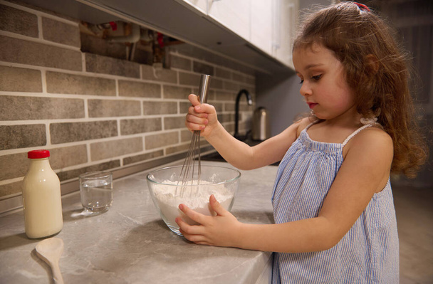 Adorable niña caucásica en vestido azul mezclando harina con ingredientes secos en un tazón de vidrio usando un batidor, preparando masa para panqueques en la cocina casera para el Martes Santo - Foto, imagen