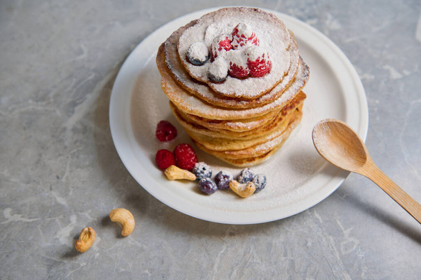 Top view of delicious homemade pancakes sprinkled with powdered sugar and decorated with raspberries, blueberries and cashews served on a white plate with a wooden spoon. Food for Shrove Tuesday - Zdjęcie, obraz
