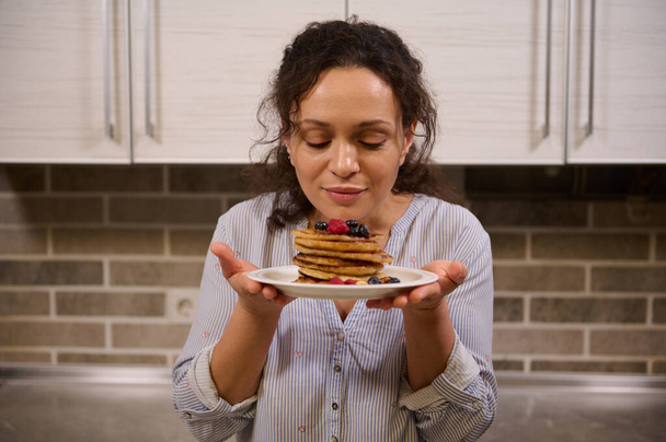 Charming woman holding a plate of delicious homemade freshly baked pancakes garnished with wild berries. Beautiful young dark-haired curly housewife with cooked pancakes for Shrovetide. Shrove Tuesday - Foto, Imagem