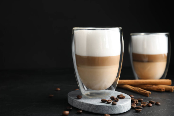 Delicious latte macchiato, cinnamon sticks and coffee beans on black table, space for text - Фото, изображение