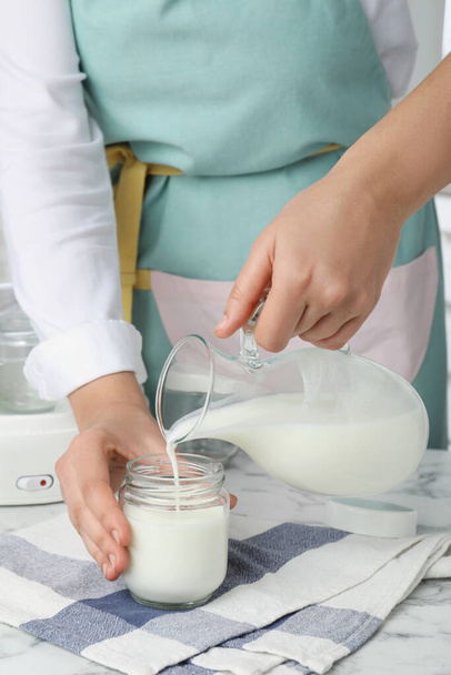 Woman pouring milk into glass jar at white marble table, closeup. Making yogurt - Foto, imagen