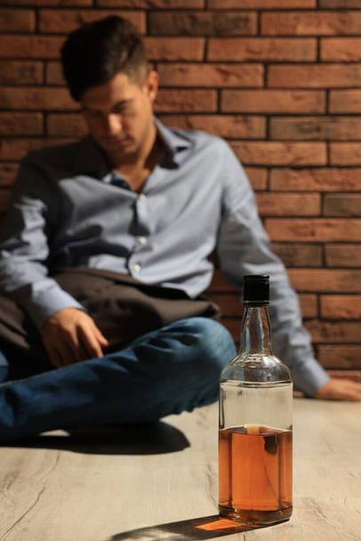 Addicted man sitting near red brick wall, focus on bottle of alcoholic drink - Fotó, kép