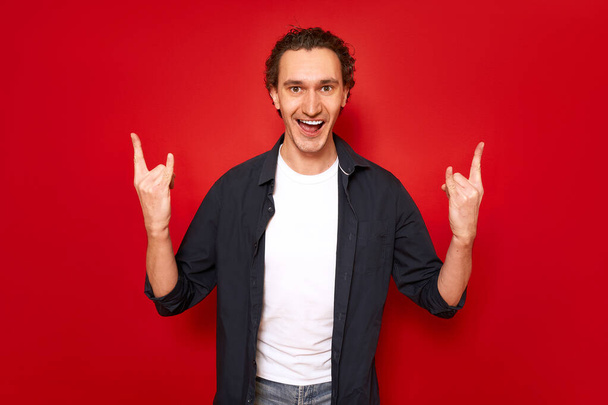 excited man with his mouth open shows rock sign with his hands depicting listening to his favorite music heavy metal. isolated on red background. concept - people, symbols, rock and roll, freedom - Photo, Image