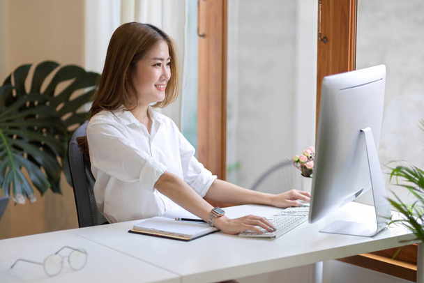 Felice asiatica giovane donna d'affari utilizzando il computer desktop nel suo moderno ufficio minimo. - Foto, immagini