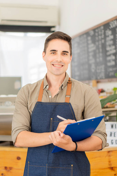 Un giovane proprietario di una caffetteria che tiene in mano un menu gastronomico si trova di fronte al negozio sorridendo felicemente. concetto di cibo e bevande - Foto, immagini