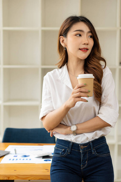 Young businesswoman, Asian woman holding a coffee cup and standing next to her desk with graph paper in the background. - Foto, afbeelding