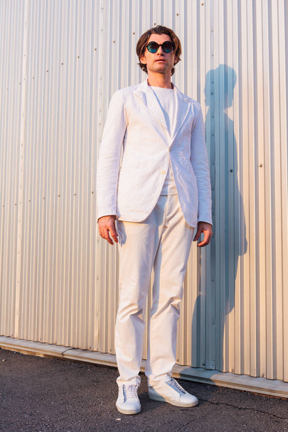Portrait of a handsome man dressed in a white suit and round hippie sunglasses standing near the wall during sunset - Φωτογραφία, εικόνα