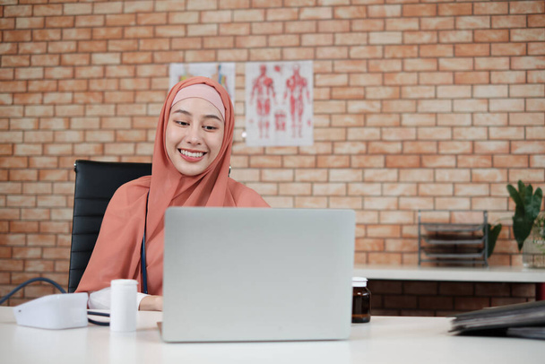Portret van een mooie vrouwelijke arts, mooie moslim in uniform met een stethoscoop, lachend en werkend met laptop in de kliniek van het ziekenhuis. Een persoon die expertise heeft in professionele behandeling. - Foto, afbeelding