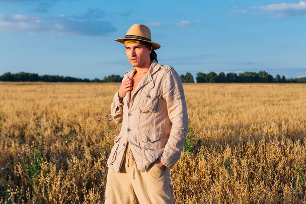 Rural Countryside. Tall handsome man dressed in a coarse linen suit and hat standing at golden oat field - Foto, immagini