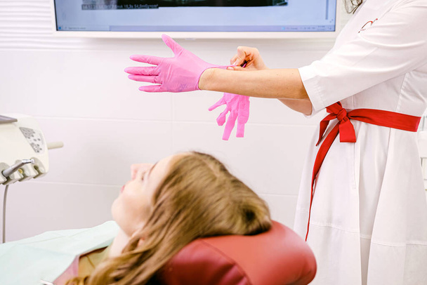 Young woman blond patient sitting on a dental chair in dentistry while woman dentist putting on gloves to start her work. - Φωτογραφία, εικόνα